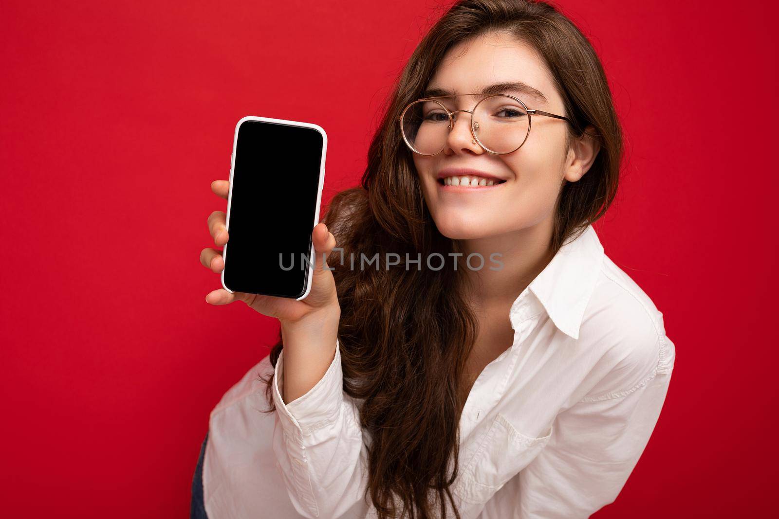 Photo of beautiful smiling young woman good looking wearing casual stylish outfit standing isolated on background with copy space holding smartphone showing phone in hand with empty screen display for mockup looking at camera.