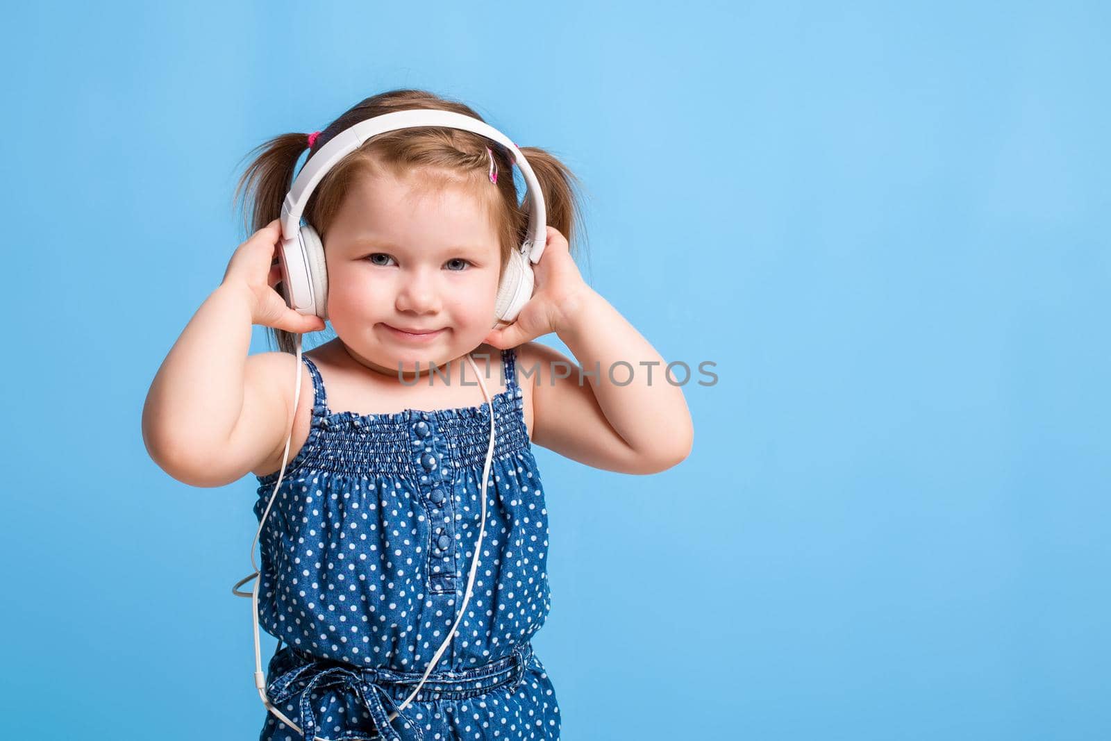 Cute little girl in headphones listening to music using a tablet and smiling on blue background. A child looks at the camera