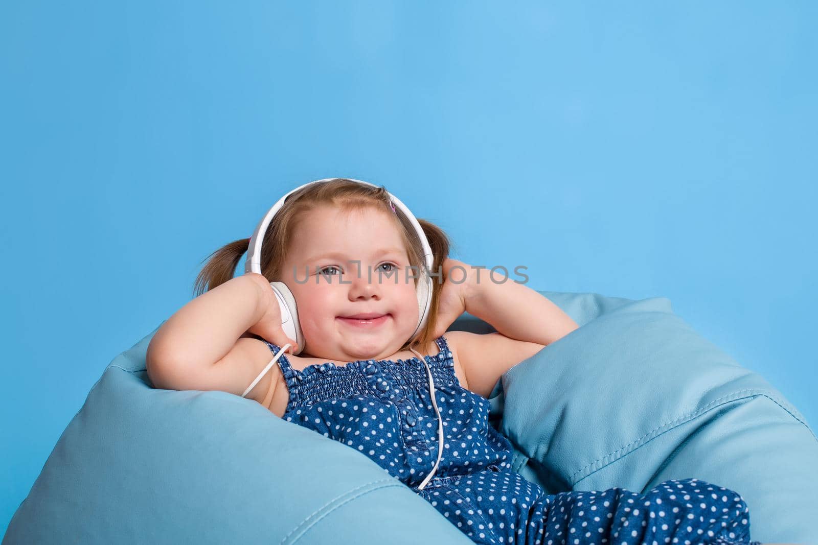 Cute little girl in headphones listening to music using a tablet and smiling while sitting on blue big bag. On blue background.