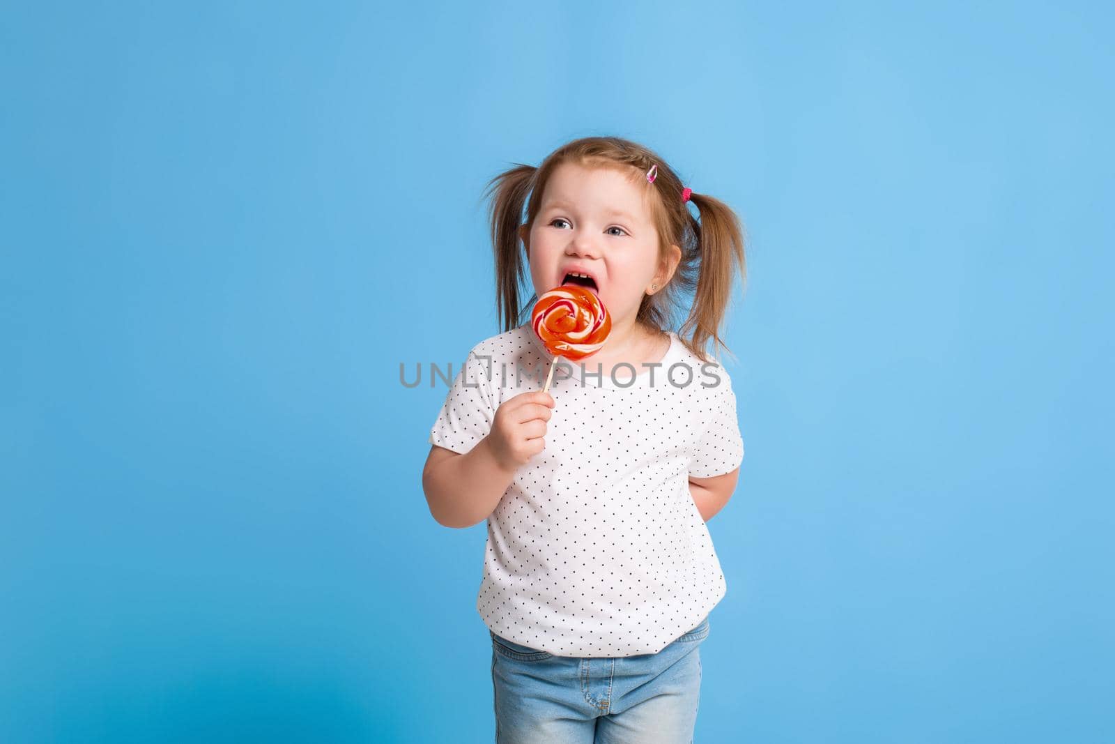 Beautiful little female child holding huge lollipop spiral candy smiling happy isolated on blue background. by nazarovsergey