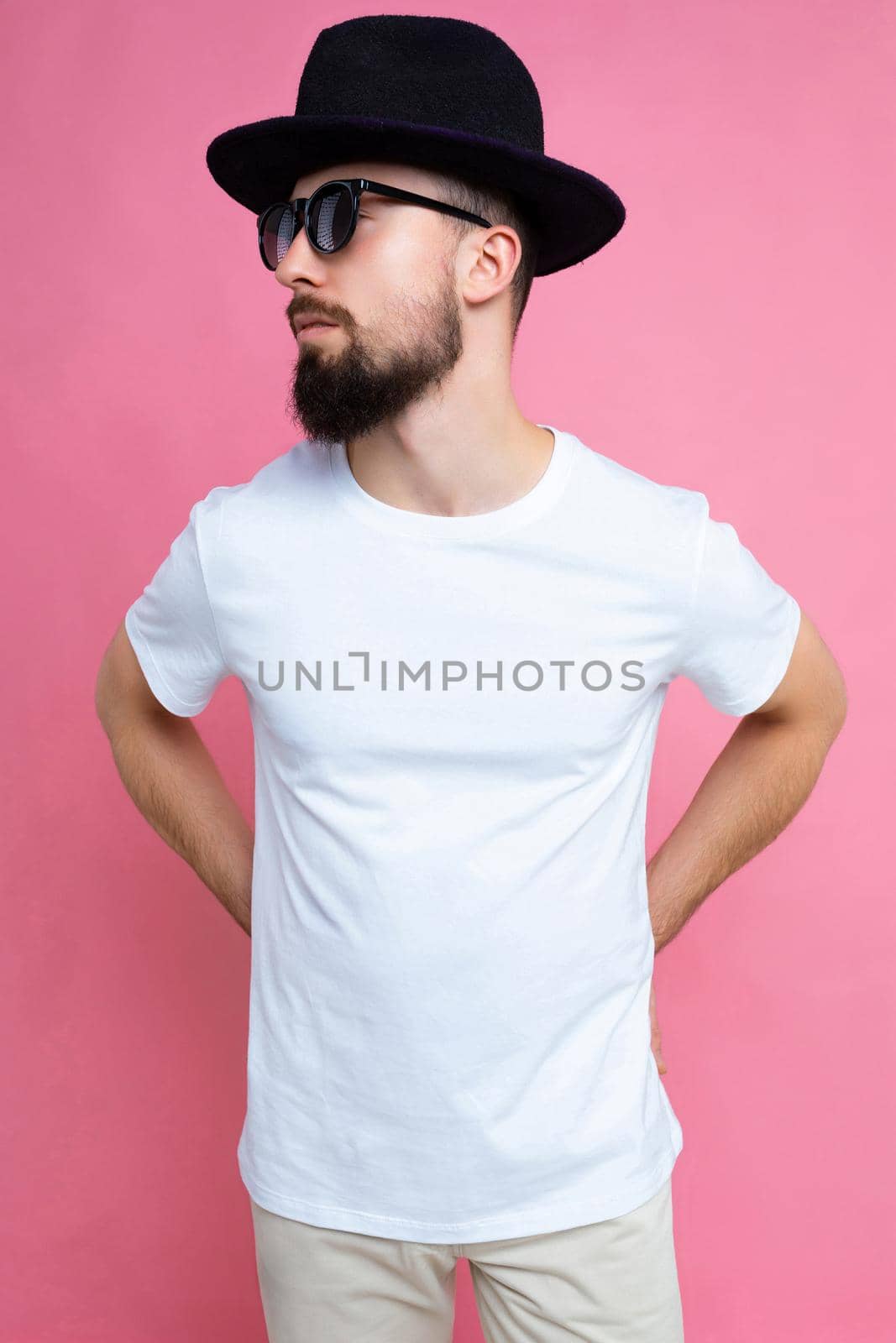 Vertical photo shot of confident brutal good looking young brunet bearded man wearing casual white t-shirt for mockup , sunglasses and stylish black hat poising isolated on pink background with empty space for text.