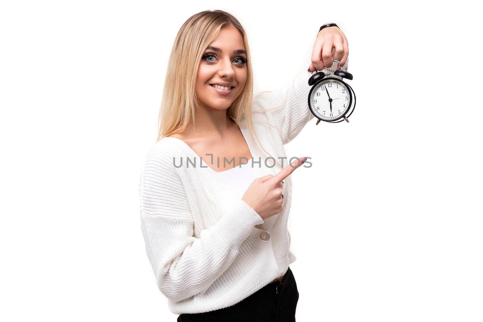 Photo of a beautiful pretty amazing positive smiling happy young blonde woman with makeup in a stylish white knitted sweater and black pants showing alarm clock isolated on a white background with copy space for text.