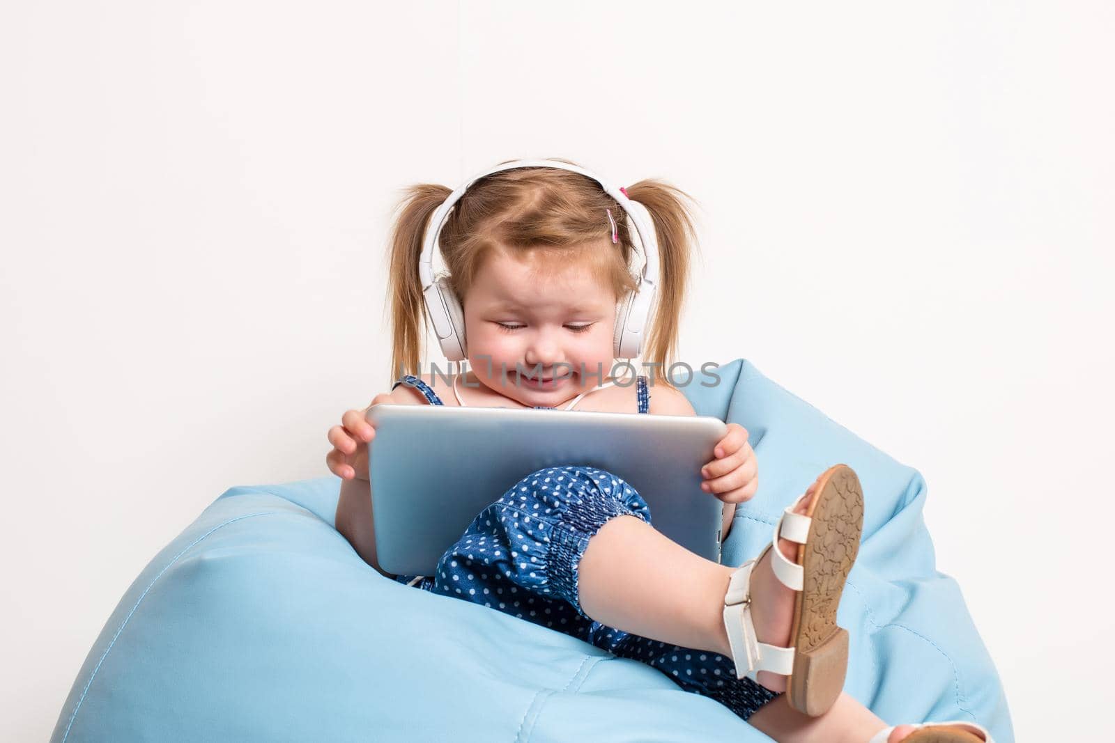 Cute little girl in headphones listening to music using a tablet and smiling while sitting on blue big bag. On white background.