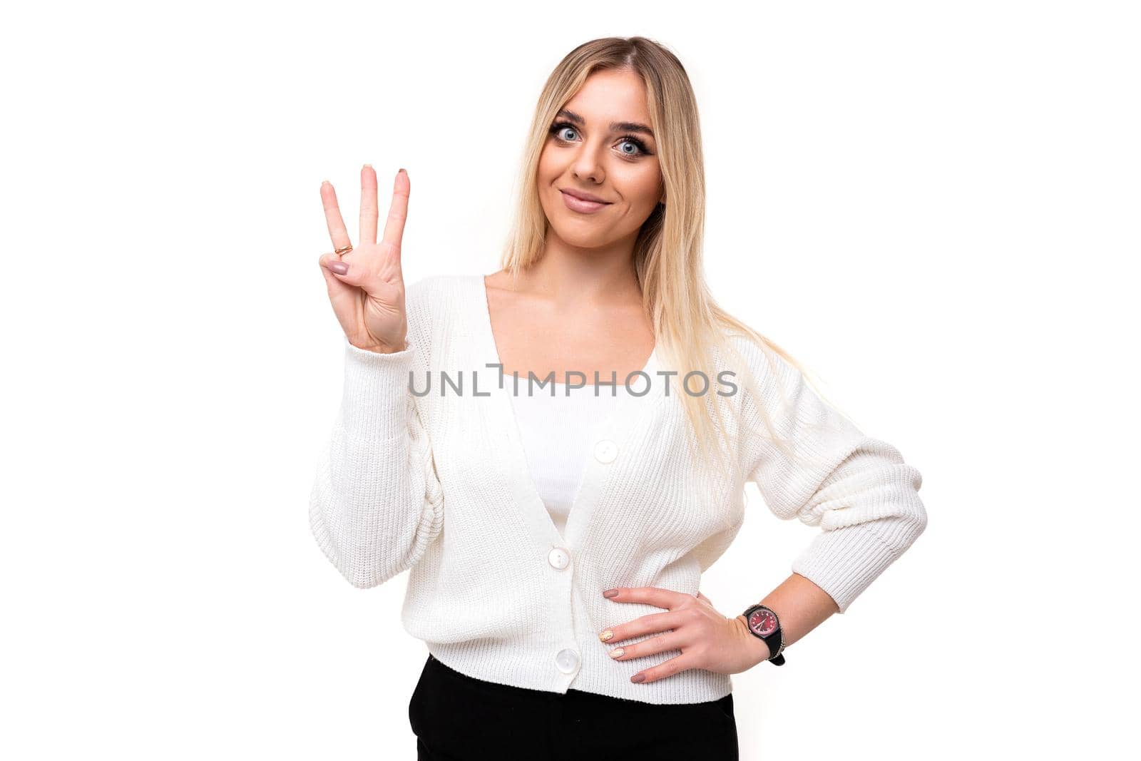 Photo of a beautiful attractive pretty amazing charming young blonde woman with makeup in a stylish white knitted sweater and black pants shows three fingers isolated on a white background with empty space for text.