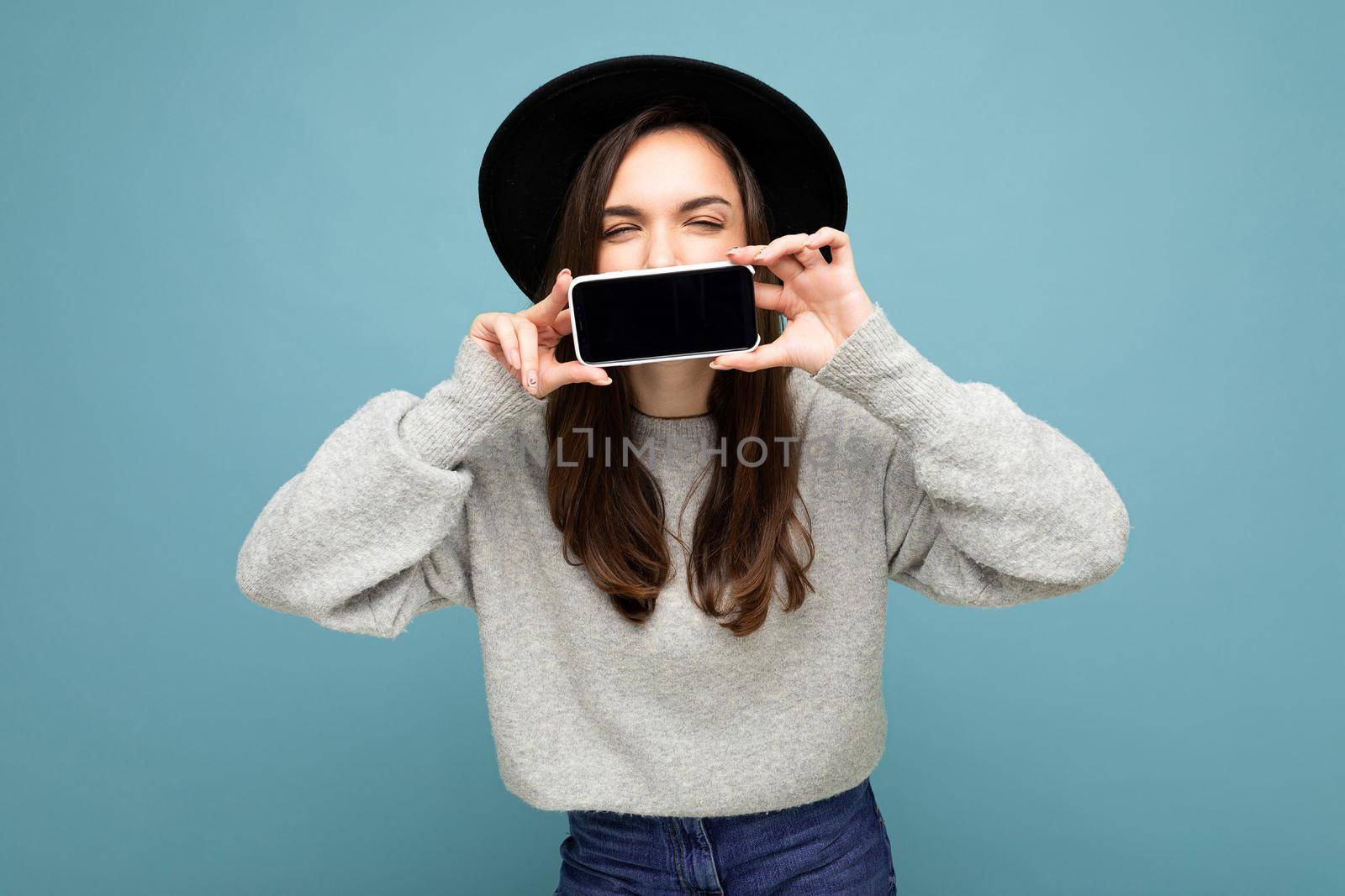 Photo of Beautiful positive woman person wearing black hat and grey sweater holding mobilephone showing smartphone isolated on background with close eyes.Mock up, cutout, free space