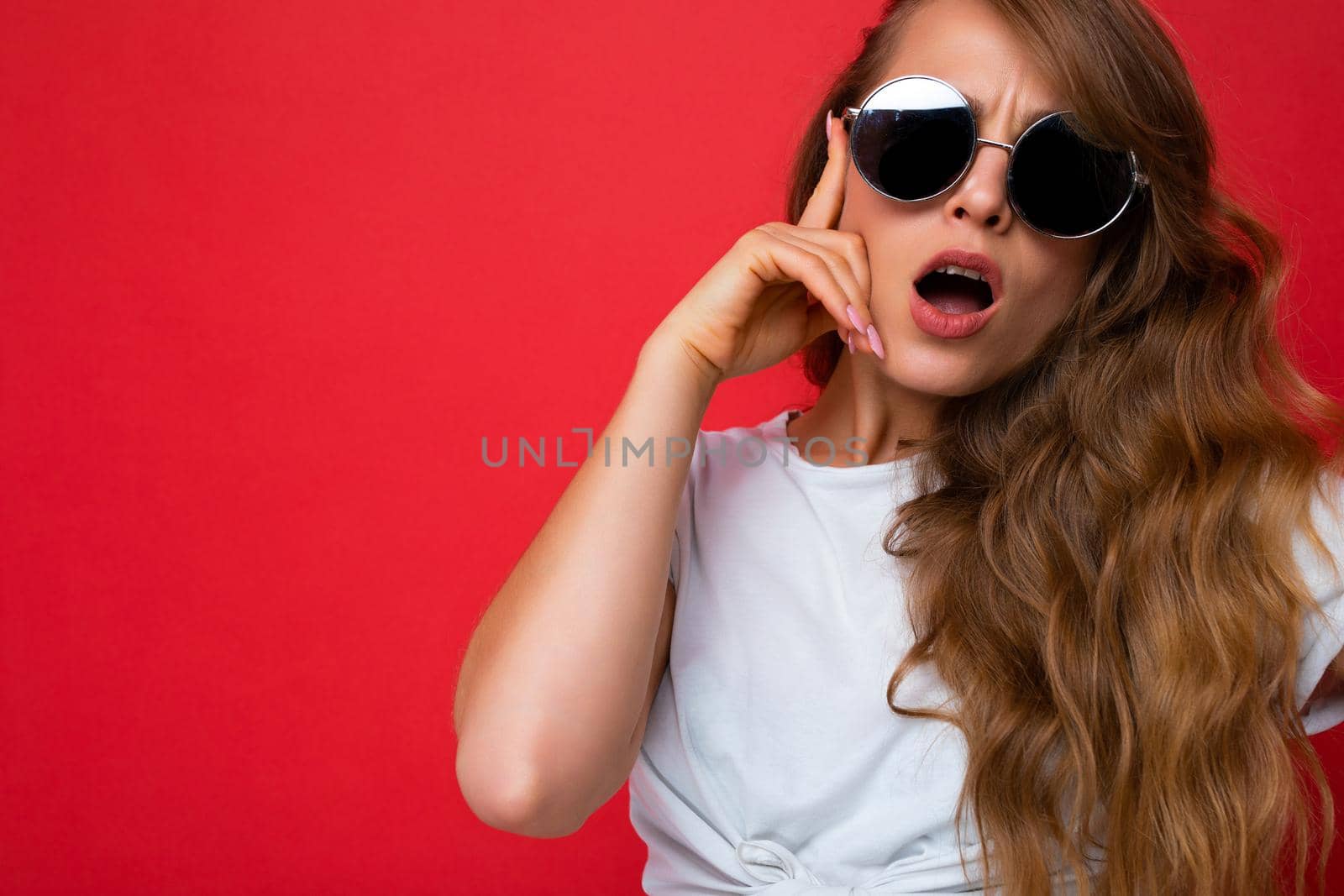 Closeup shot of beautiful surprised young dark blonde curly woman isolated over red background wall wearing casual white t-shirt and stylish sunglasses looking at camera with open mouth. empty space