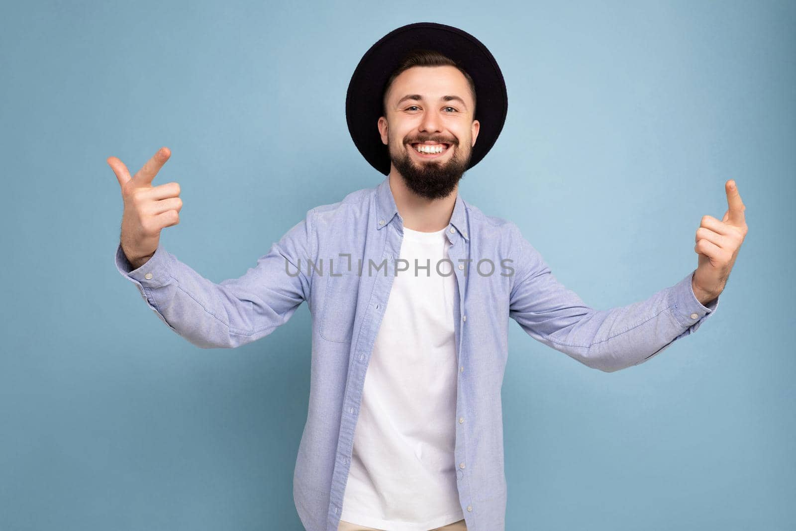 Photo shot of positive happy smiling good looking young brunet bearded man wearing casual blue shirt and white t-shirt and stylish black hat poising isolated on blue background with free space for text.