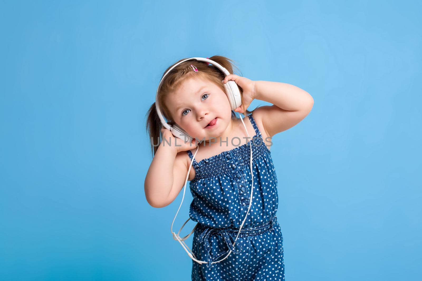 Cute little girl in headphones listening to music using a tablet and smiling on blue background. A child looks at the camera