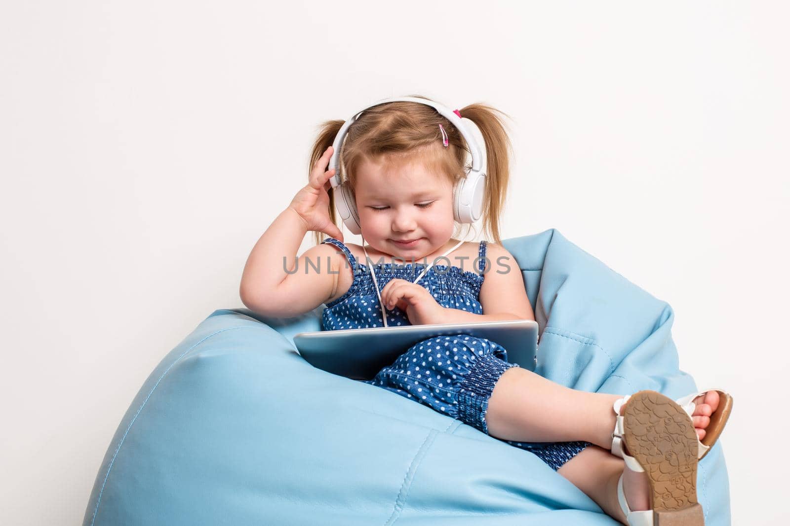 Cute little girl in headphones listening to music using a tablet and smiling while sitting on blue big bag by nazarovsergey