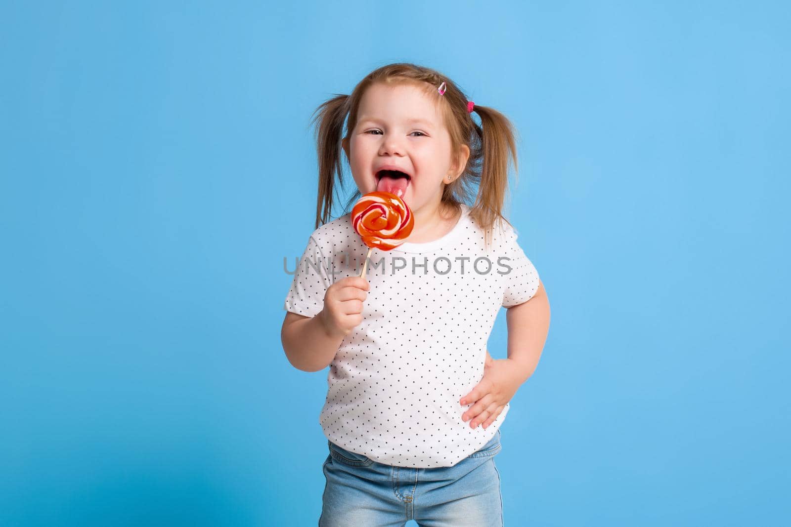 Beautiful little female child holding huge lollipop spiral candy smiling happy isolated on blue background. In children loving sweet and caramel concept