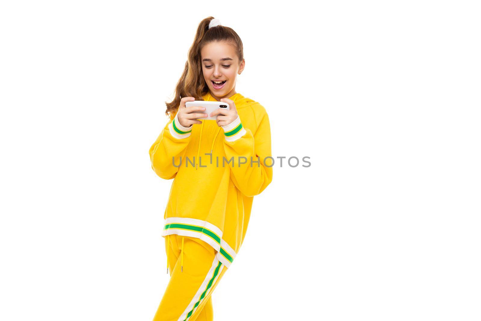 Photo of a beautiful smiling cheerful cute girl with dark hair with a ponytail in a stylish yellow tracksuit with green stripes plays a mobile phone with a white case isolated on a white background with free space for text.