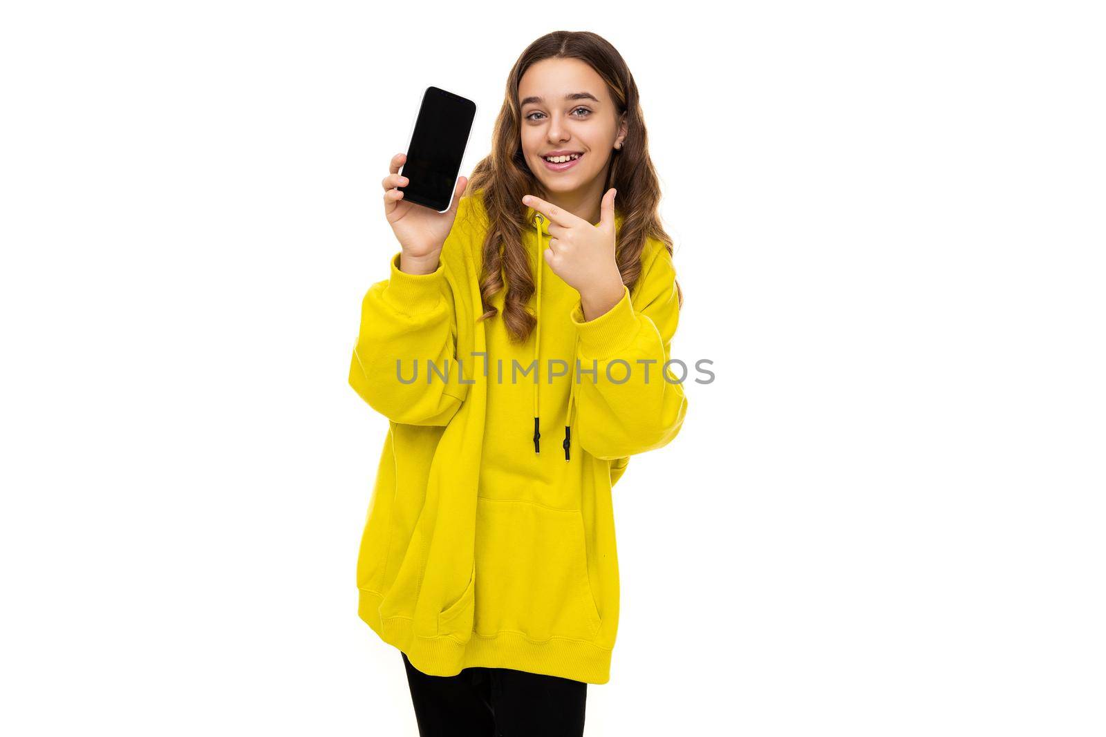 Photo of a beautiful adorable smiling positive funny brunette teenage girl in a stylish yellow sports hoodie holding a mobile phone with a black blank screen isolated on a white background with free space for text.