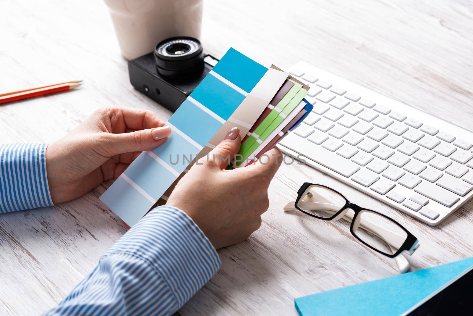 Interior designer holding color swatches at wooden desk. Office workplace with computer keyboard and camera. Brainstorming and creativity. Selection of color palette for web or product design.