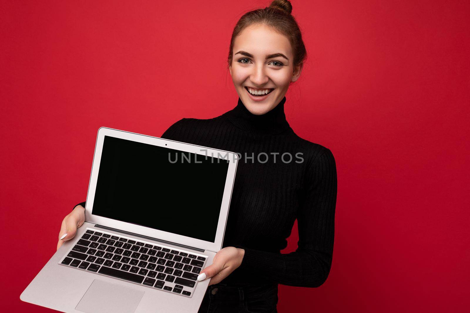 Photo of beautiful young woman holding computer laptop looking at camera isolated over colourful background.