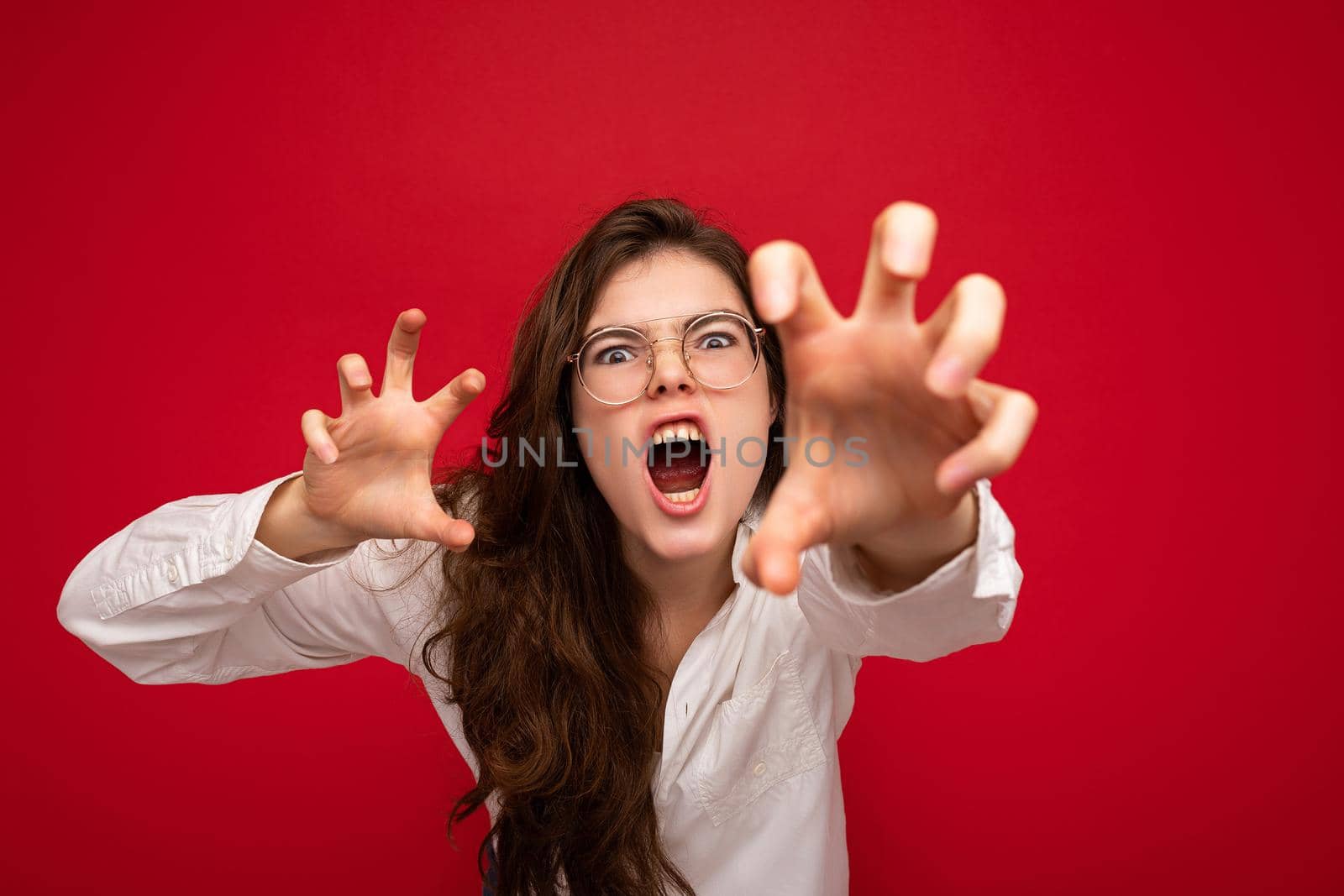 Portrait of young emotional attractive brunette woman with sincere emotions wearing casual white shirt and optical glasses isolated on red background with free space and doing claw gesture as cat with agressive and sexy expression. Roar concept.