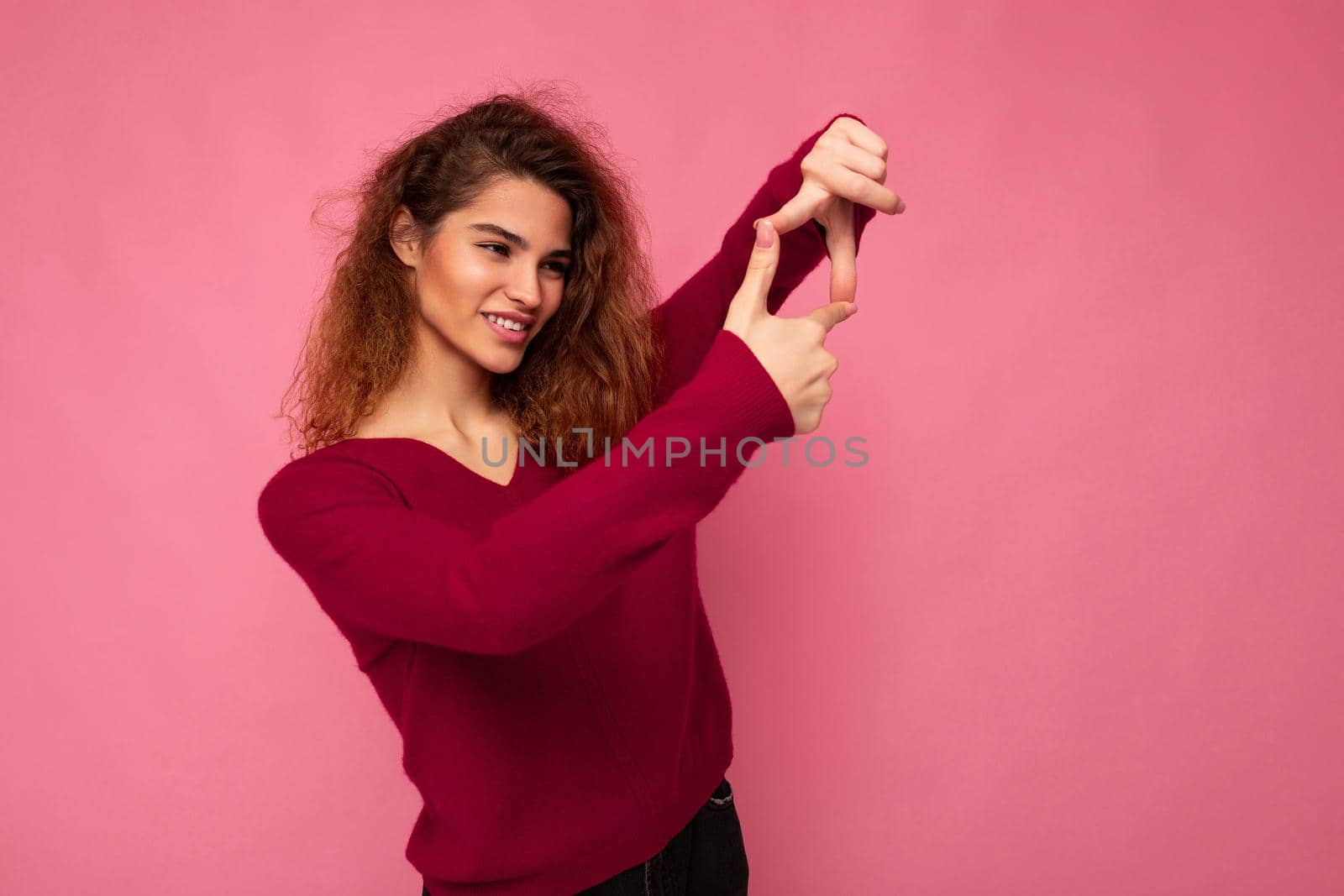 Portrait of young positive pretty brunette curly woman with sincere emotions wearing trendy pink pullover isolated on pink background with copy space and showing camera gesture with hands taking photo.