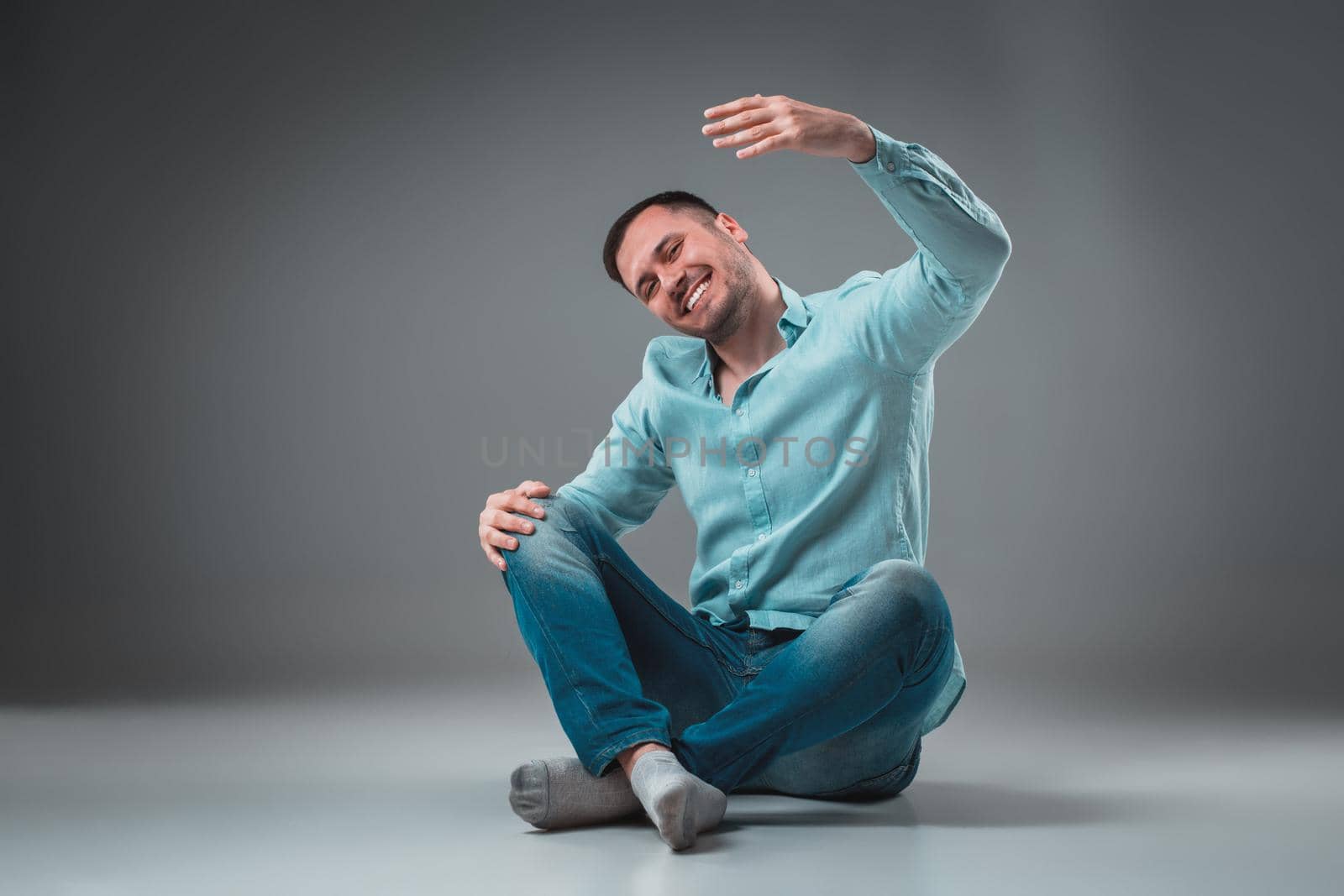 Handsome young man sitting on a floor with raised hands, isolated on gray background. Emotion concept