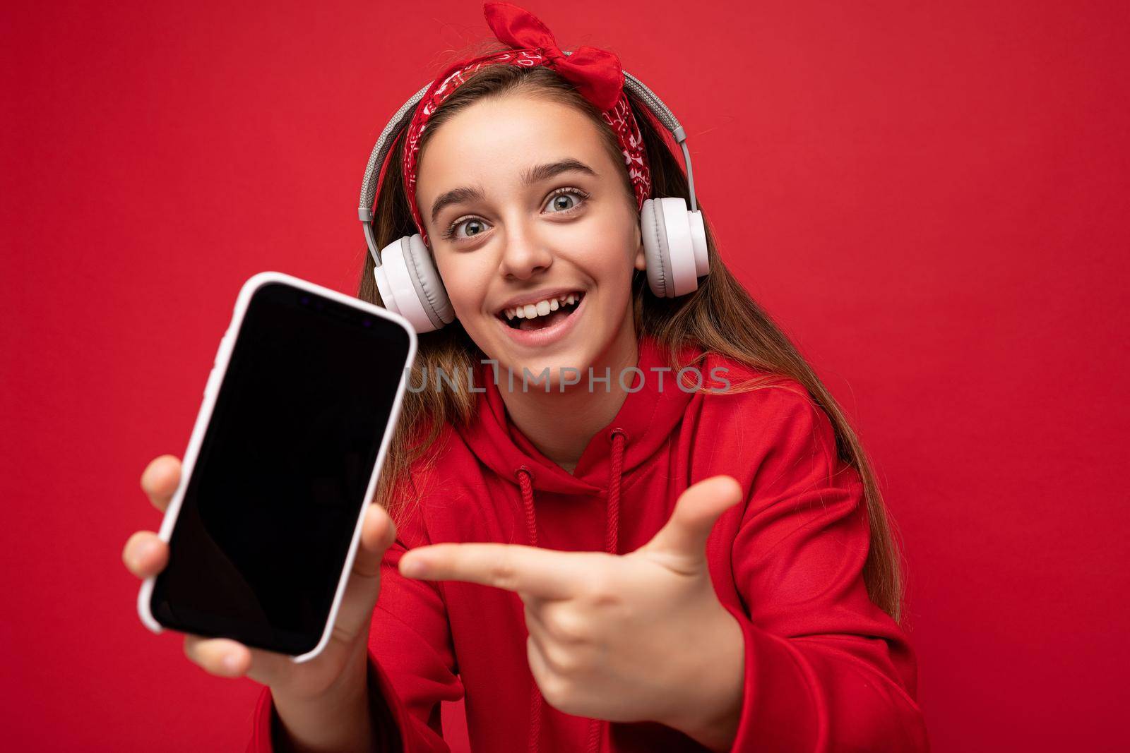 Closeup Photo of positive smiling surprised cute brunette girl wearing red hoodie isolated on red background holding and showing smartphone with empty display for cutout wearing white wireless headphones listening to funny music looking at camera and pointing finger at gadjet.