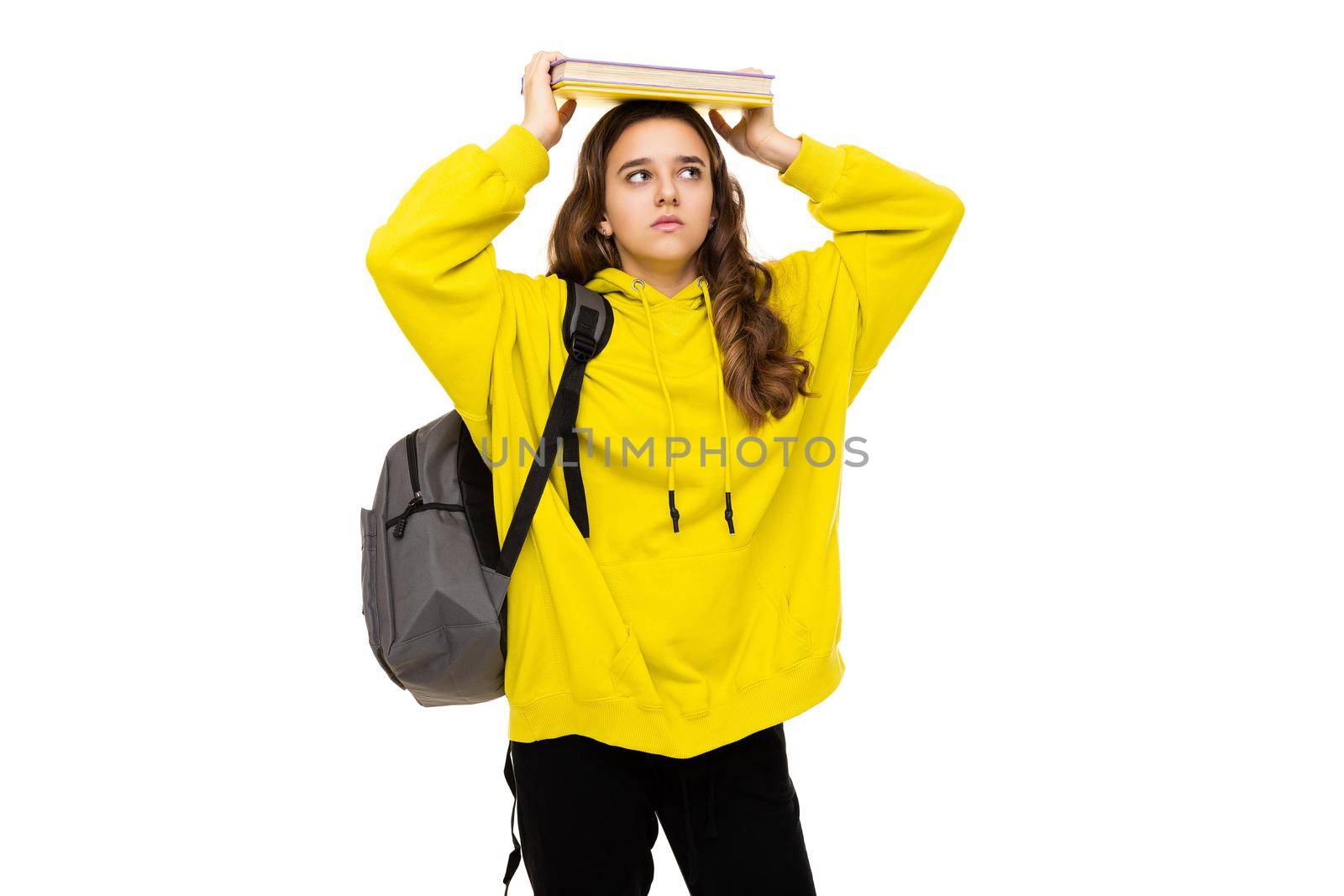 Photo of a beautiful brunette teenage girl in a stylish yellow sports hoodie with a gray backpack holding a lot of books to study with her head isolated on a white background with free space for text. School concept by TRMK
