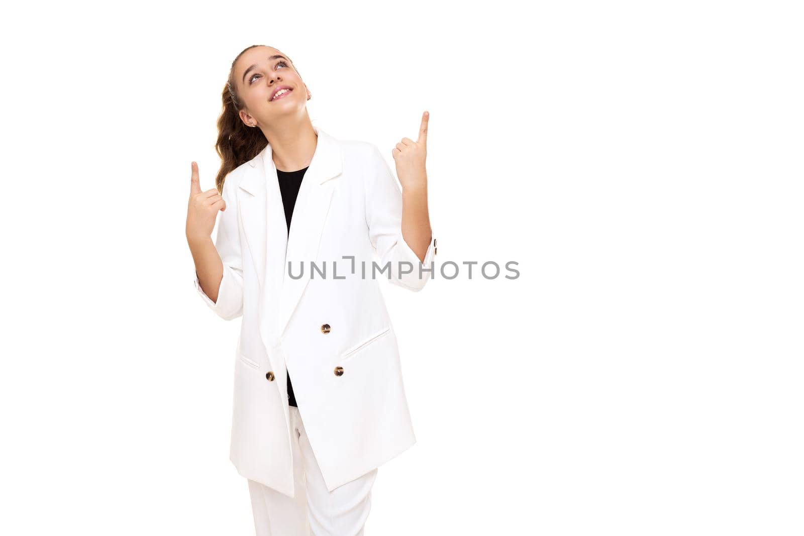 Photo of a beautiful cute adorable smiling brunette teenage girl with a ponytail in a stylish white jacket and white pants isolated on a white background shows a fingers up on a free space for text.