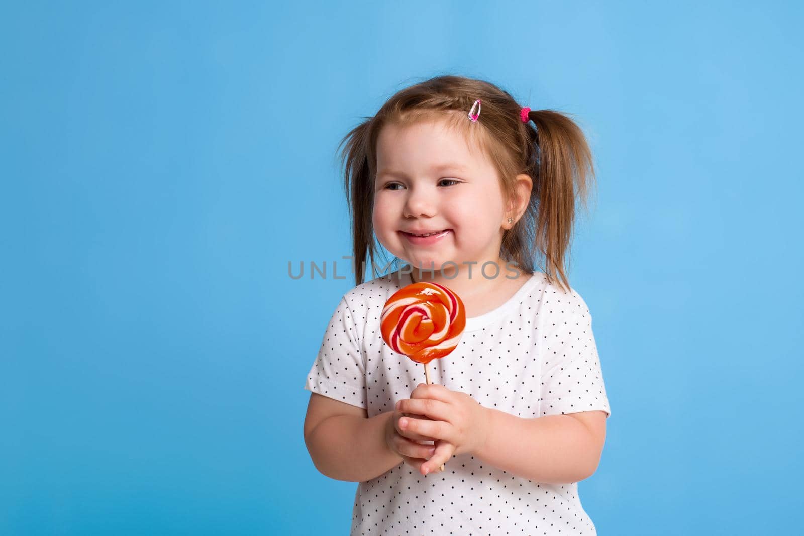 Beautiful little female child holding huge lollipop spiral candy smiling happy isolated on blue background. by nazarovsergey