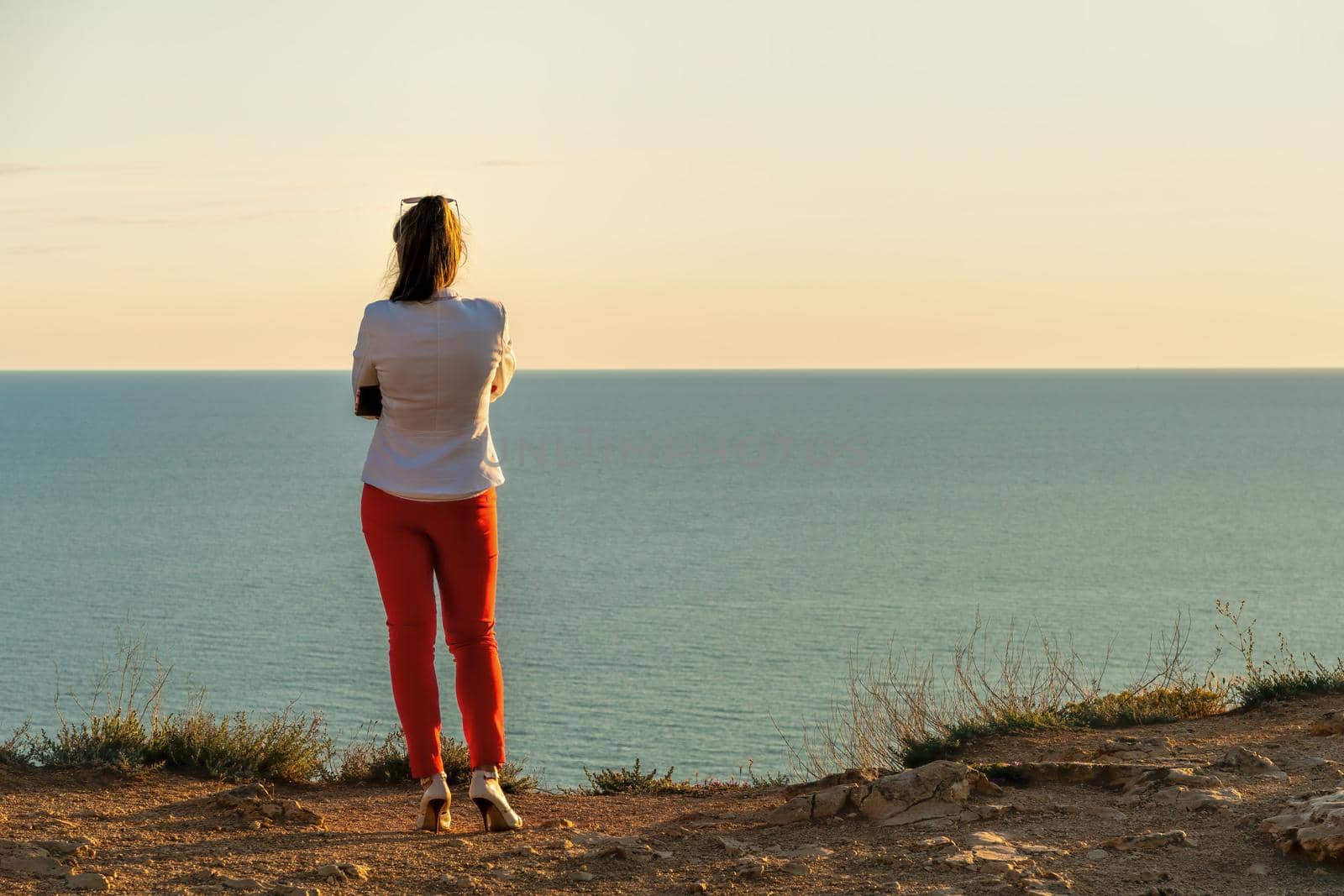 Young beautiful brunete woman in a white jacket and red pants, standing on a cliff over the sea and wait for a call. back view. Copy space. The concept of calmness, silence and unity with nature