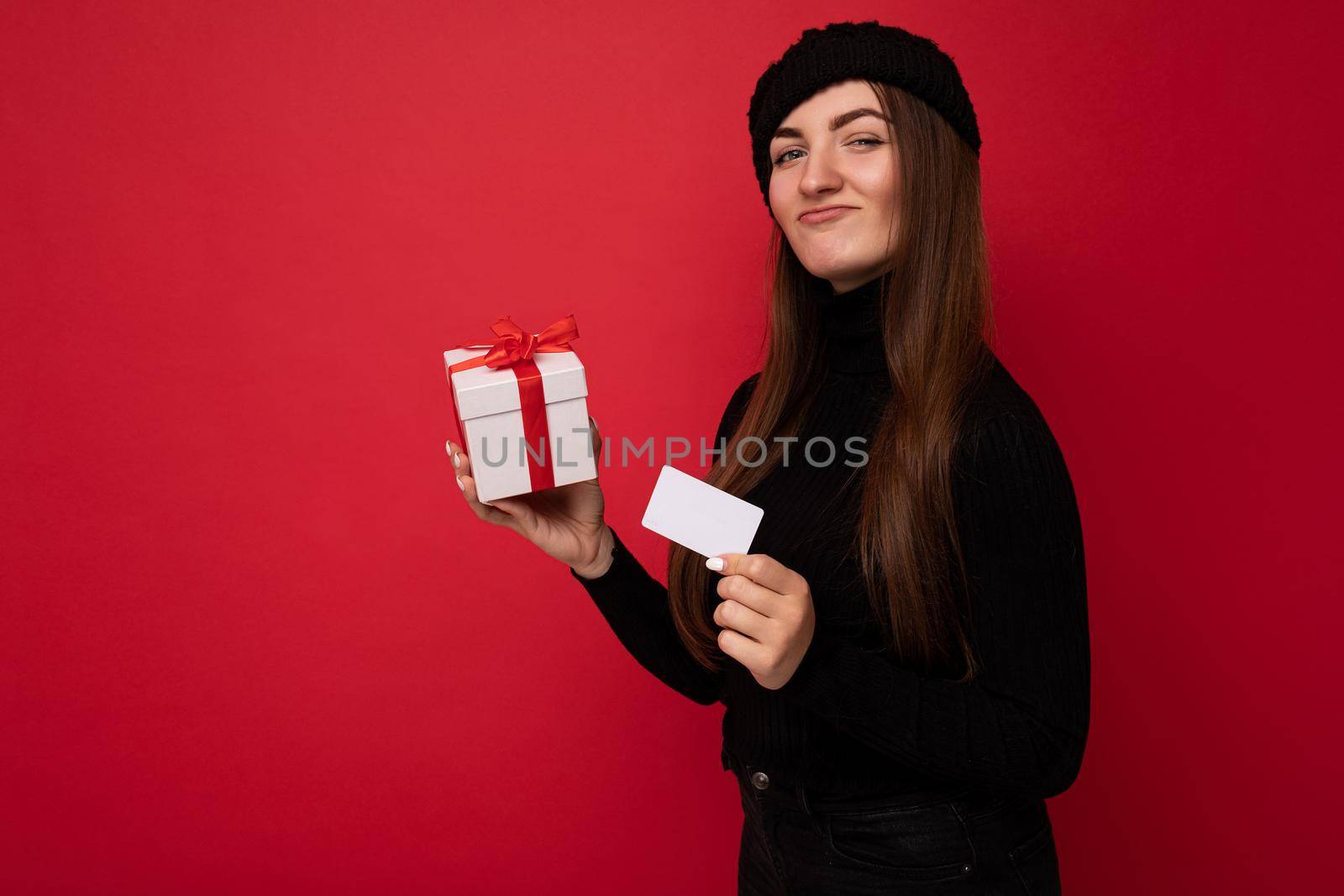 Attractive positive young brunette woman wearing black sweater and hat isolated on red background holding credit card and white gift box with red ribbon looking at camera.