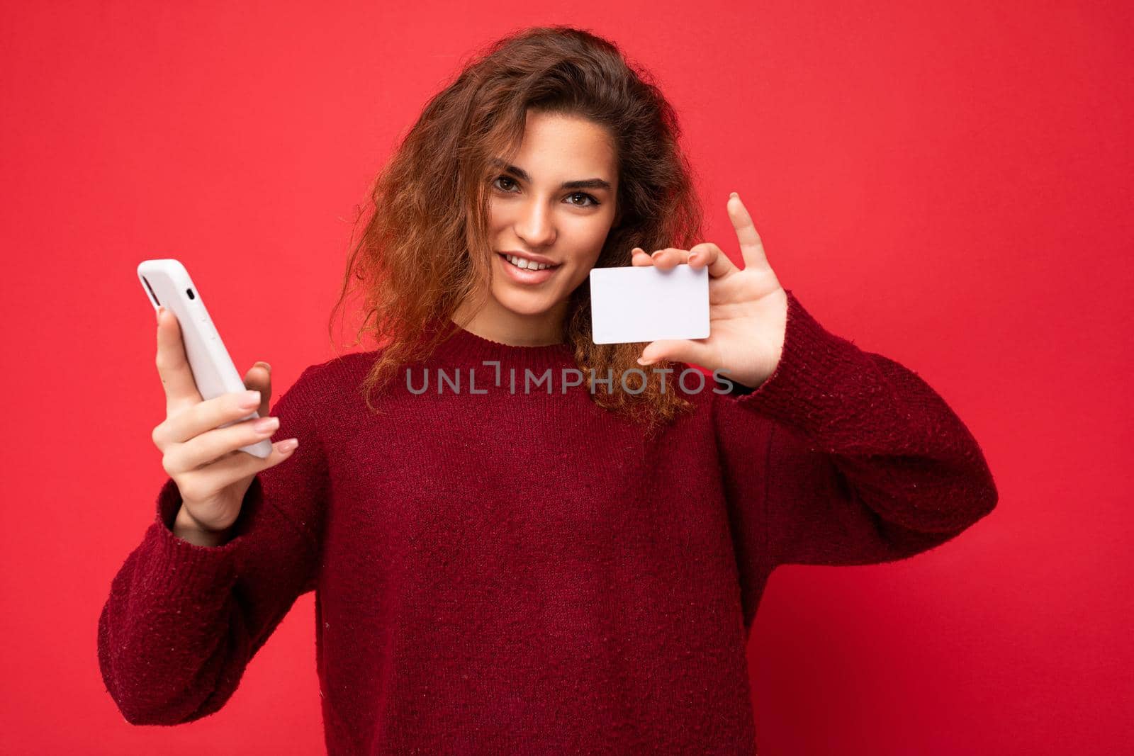 Young smiling beautiful curly brunette woman with sincere emotions poising isolated over background wall with empty space wearing casual dark red sweater holding credit card and using mobile phone . Internet banking concept.