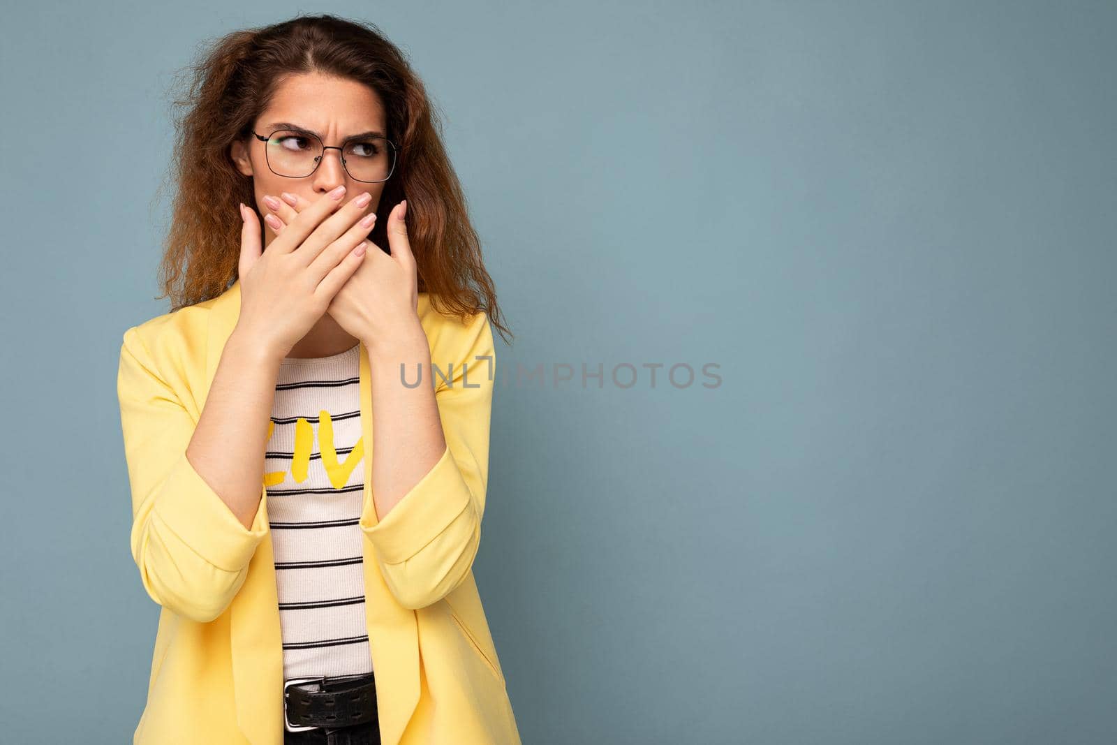 Photo shot of young angry sad nice cute brunette curly woman with sincere emotions wearing trendy yellow jacket isolated on blue background with copy space and covering mouth.