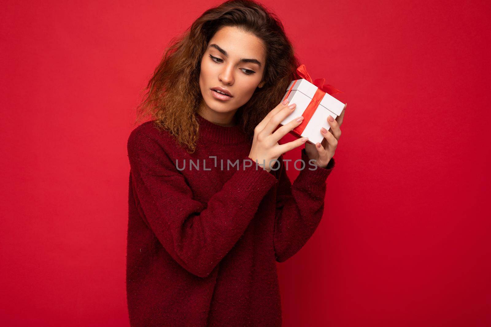 Attractive positive concentrated young brunet curly woman isolated over red background wall wearing red sweater holding gift box and trying guess the surprise.