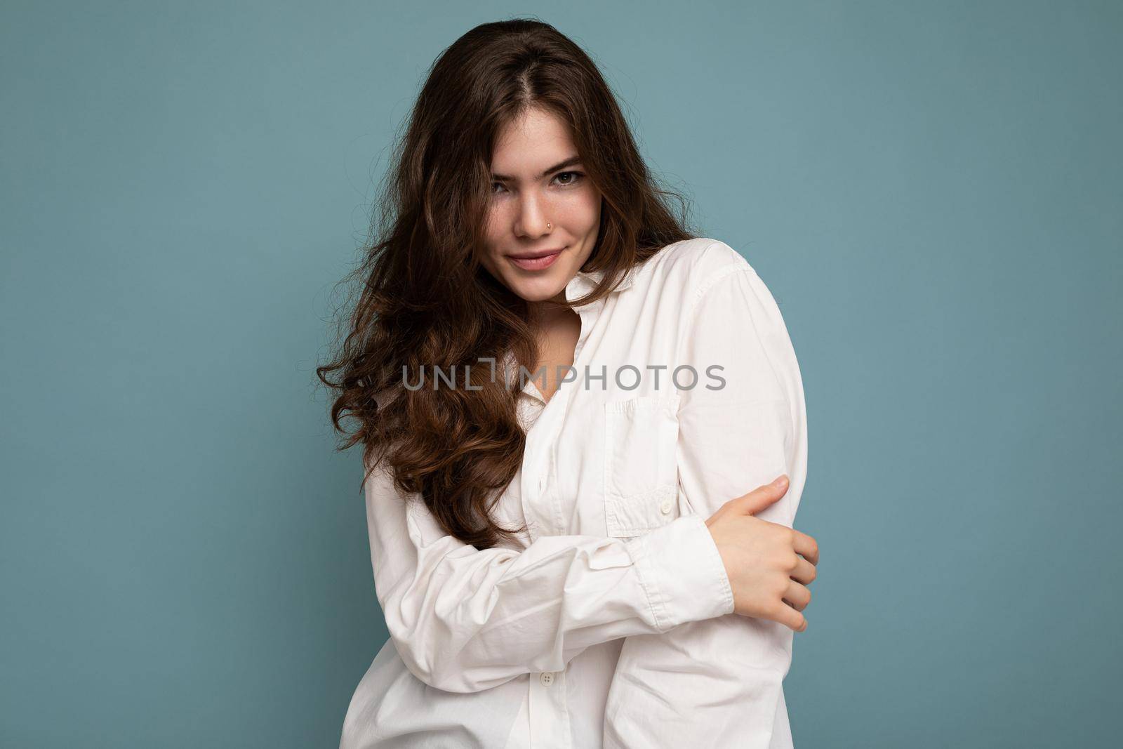 Portrait of young cute tender attractive winsome charming curly brunette woman with sincere emotions wearing stylish white shirt isolated on blue background with copy space.