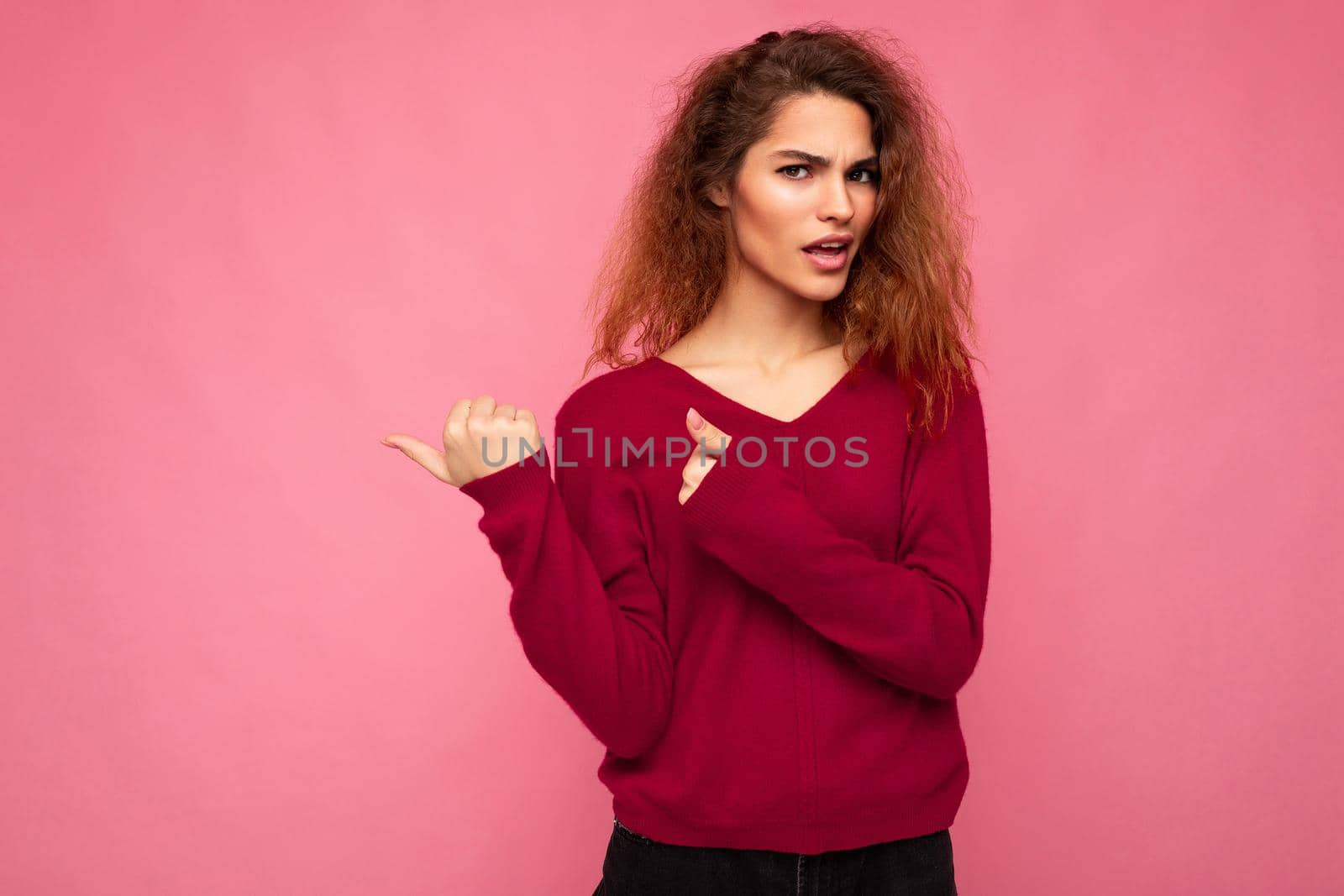 Photo of young shocked beautiful brunette wavy woman with sincere emotions wearing casual pink jersey isolated over pink background with copy space and pointing at free space for advertising.