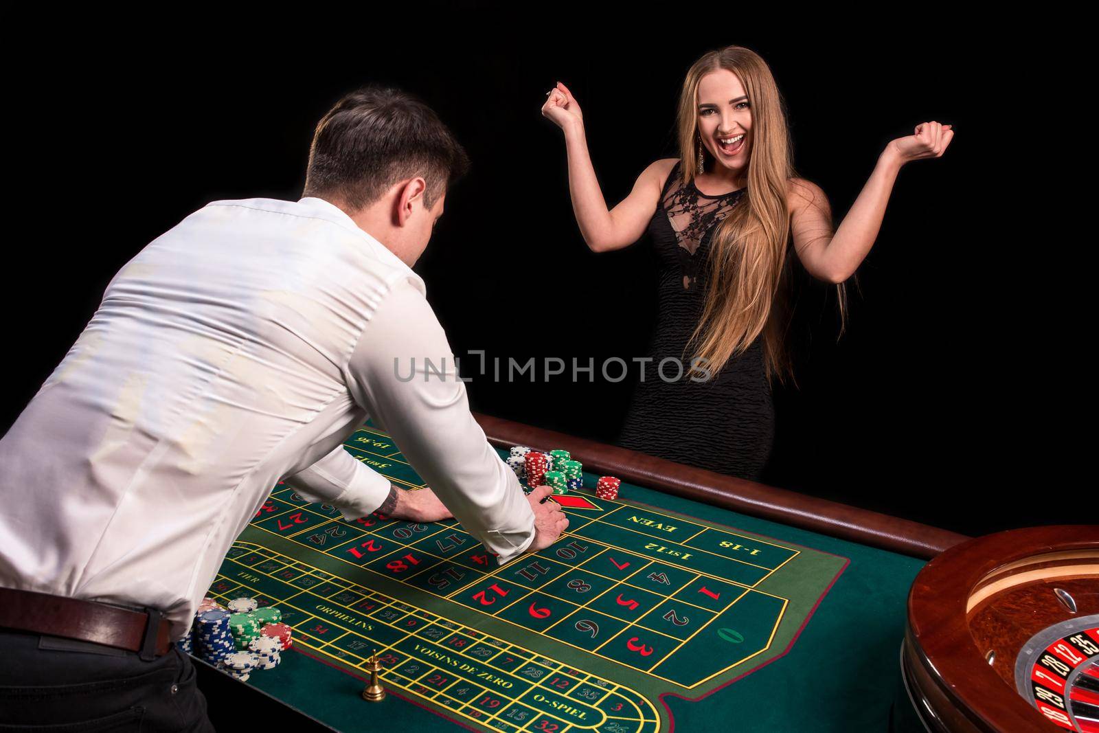 A close-up on the back of the croupier in a white shirt, image of green casino table with roulette and chips, a rich woman betting of gambling in the background by nazarovsergey