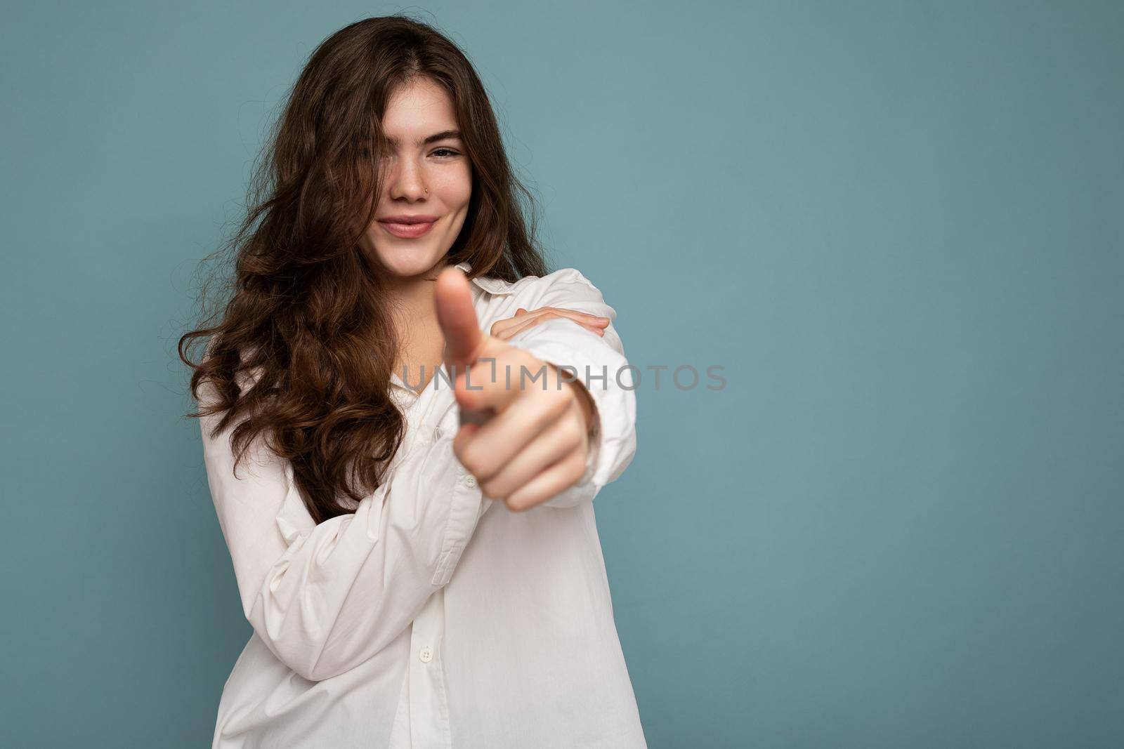 Portrait of young positive happy attractive curly brunette woman with sincere emotions wearing casual white shirt isolated on blue background with empty space and pointing finger at you.