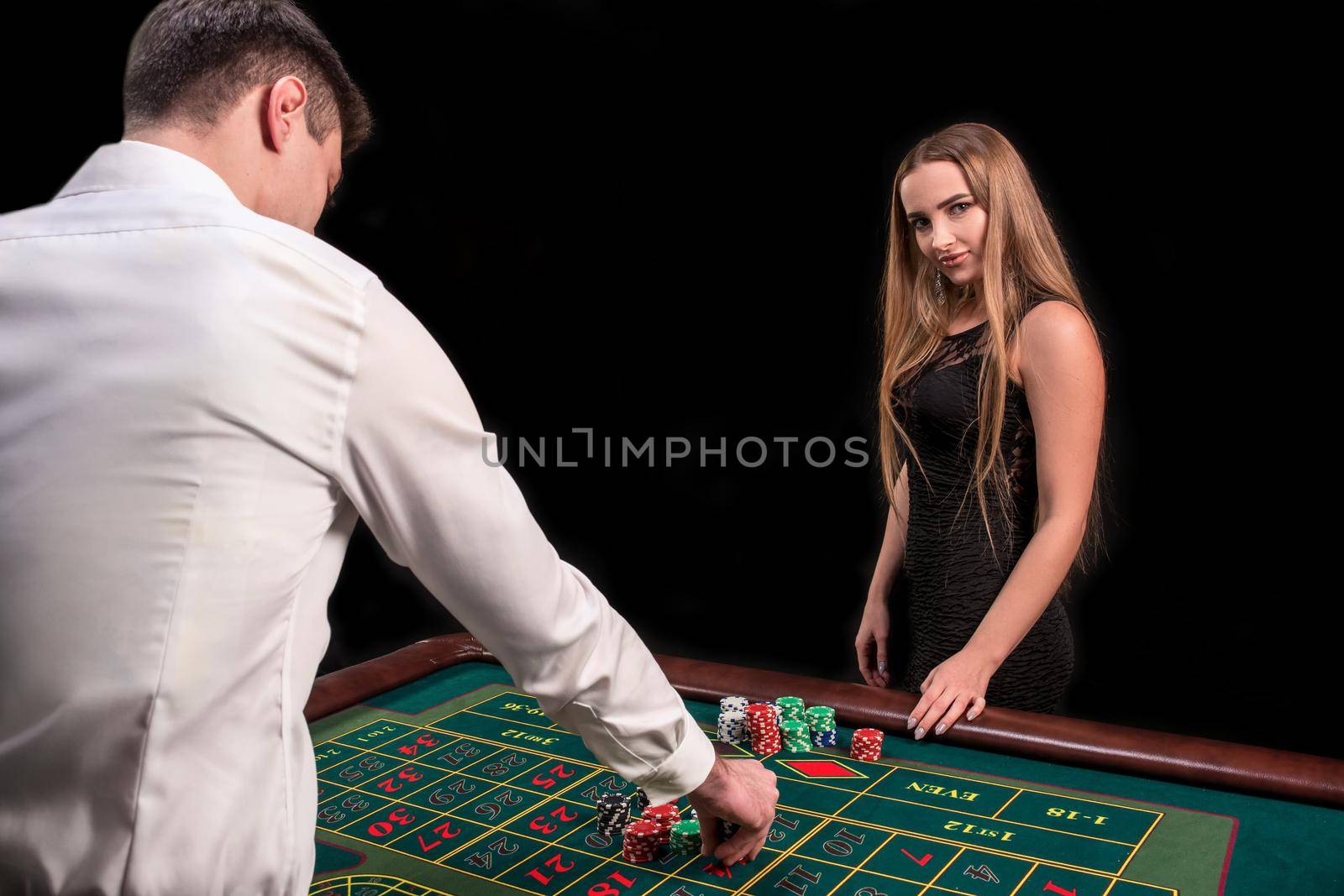 A close-up on the back of the croupier in a white shirt, image of green casino table with roulette and chips, a rich woman betting of gambling in the background. Casino. Gambling. Roulette. Betting