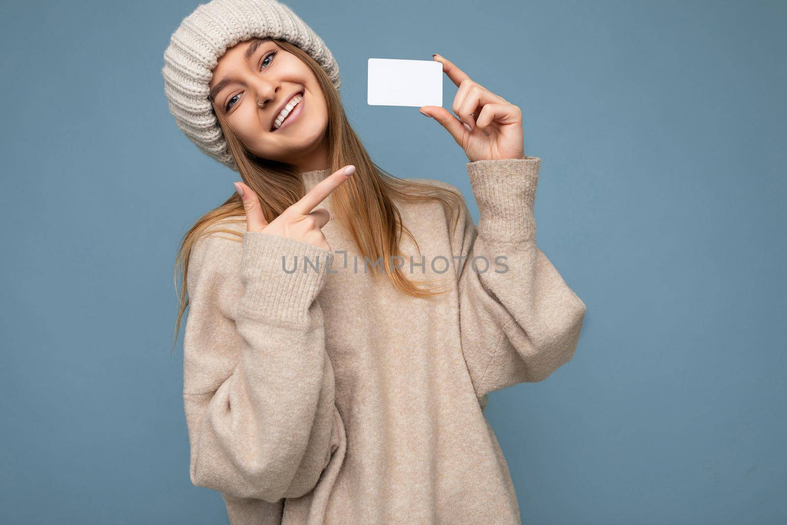 Photo of attractive pretty beautiful positive happy smiling young dark blond woman isolated over blue background with empty space wearing beige warm sweater and knitted beige hat and showing at plastic credit card for mockup.