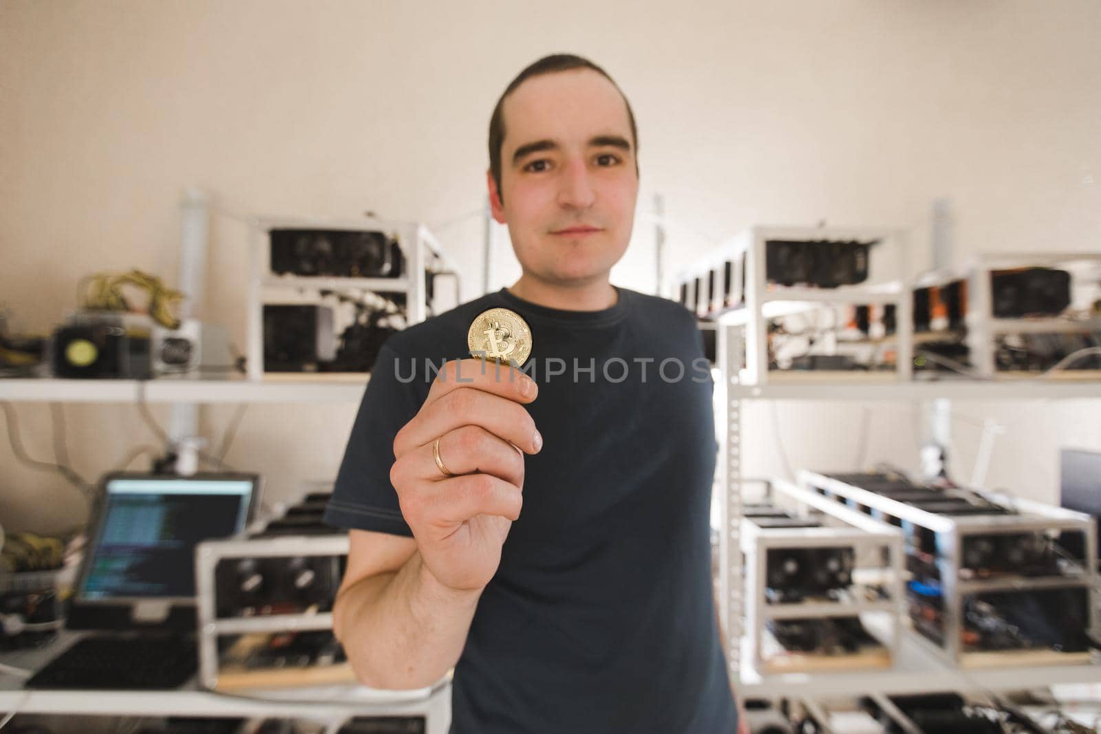 Confident man in dark clothes shows bitcoin against the background of computer devices