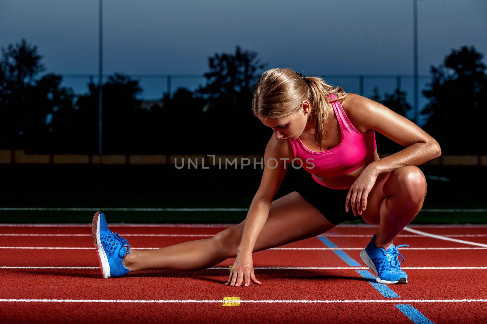 Attractive young woman athlete stretching legs on stadium. The end of training at sunset.