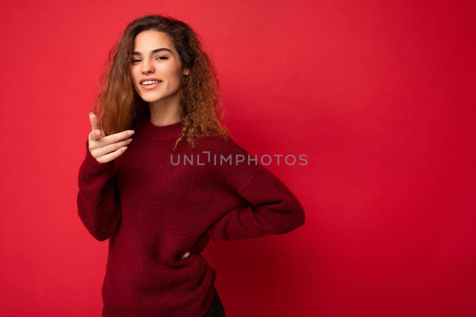 Young positive happy beautiful brunette curly woman with sincere emotions wearing casual red pullover isolated on red background with copy space.