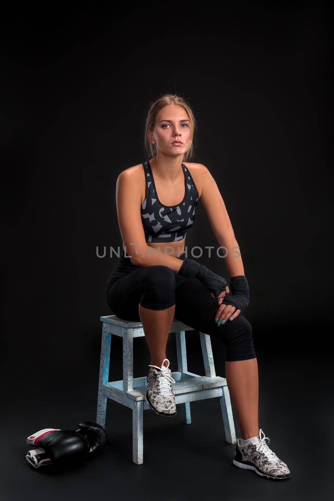 Young fit woman looking at the camera while wrapping her arms with bandage tape. Attractive girl preparing for the kickboxing training. Woman in black sports uniform on a black background