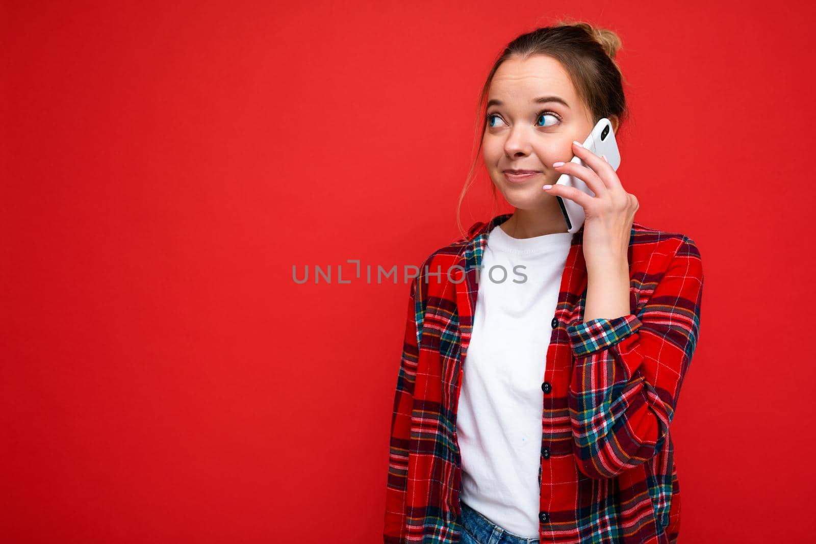 Photo of young attractive beautiful positive amazed blonde woman in stylish red shirt. Sexy carefree female person isolated on red wall background in studio with empty space for text and communicating on mobile phone with friend.