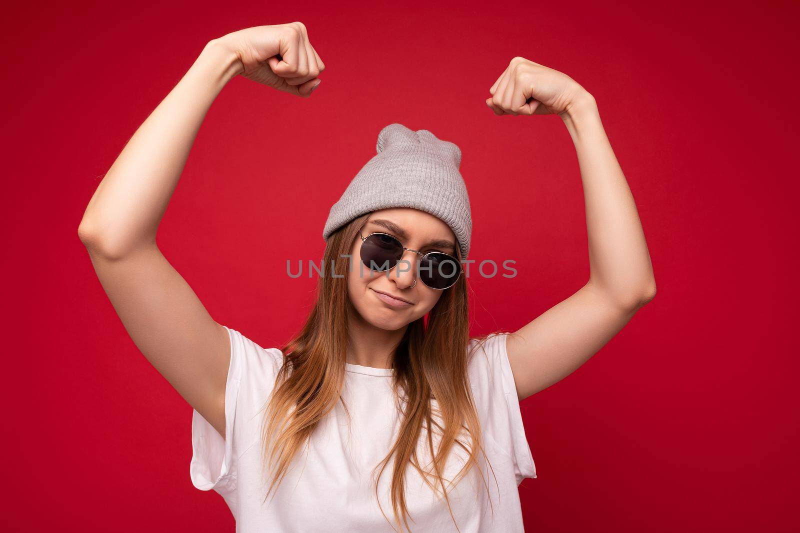 Portrait of young emotional positive funny beautiful dark blond woman wearing casual white t-shirt with empty space for mock up gray hat and sunglasses isolated over red background with free space. Female model showing powerful muscles and biceps. Fitness and workout concept.