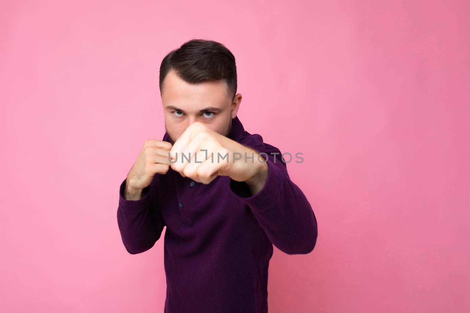 Photo shot of handsome self-confident and good looking young brunet bearded man wearing casual and stylish purple longsleeve poising isolated on pink background with empty space for text. Ready to fight with fist.