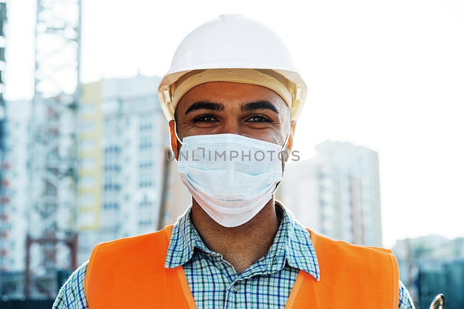Portrait of mixed race man builder in workwear and hardhat wearing medical mask, close up photo