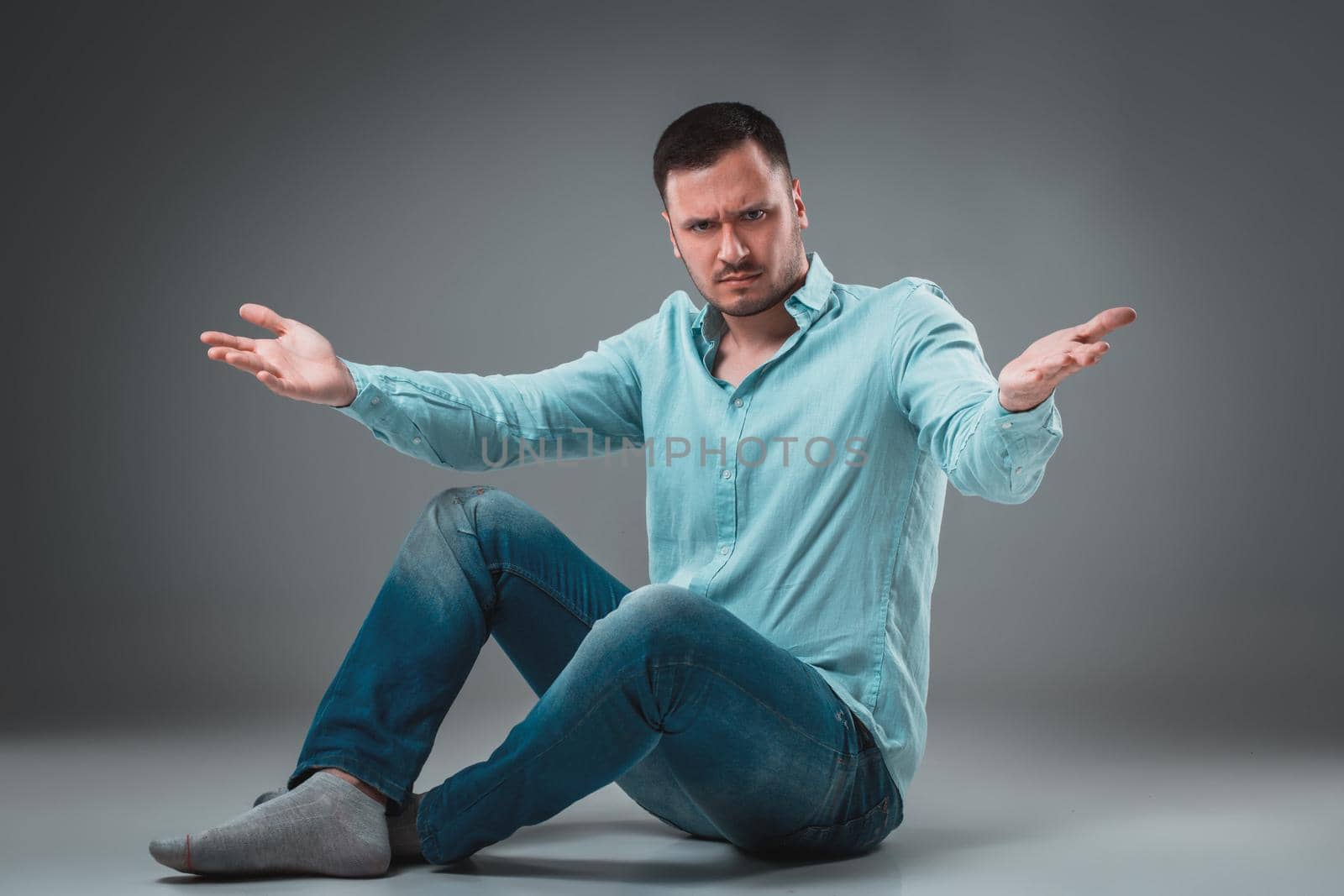 Handsome young man sitting on a floor with raised hands gesturing on gray background. A man in jeans and a blue shirt