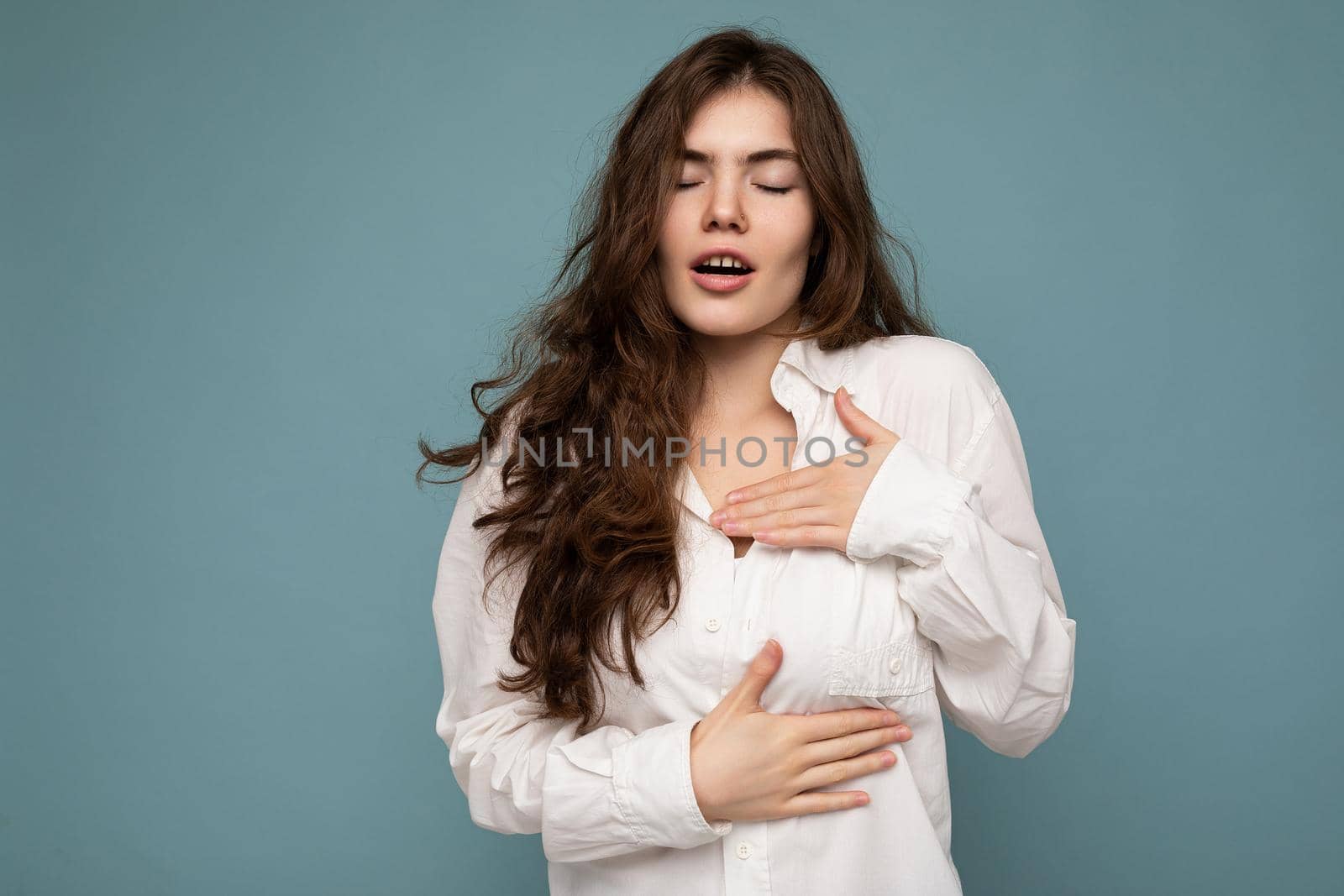 Portrait of young emotional amazed beautiful wave-haired brunette woman with sincere emotions wearing casual white shirt isolated on blue background with empty space and touches chest with hands.