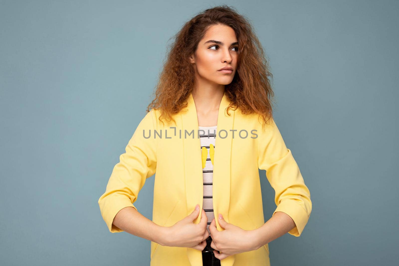 Young self-confident serious beautiful curly brunette woman with sincere emotions poising isolated over background wall with empty space wearing casual yellow jacket. Feelings concept.