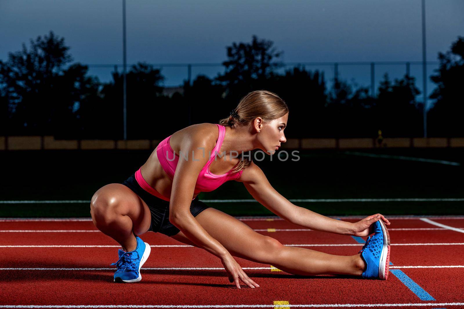 Attractive young woman athlete stretching legs on stadium. The end of training at sunset.