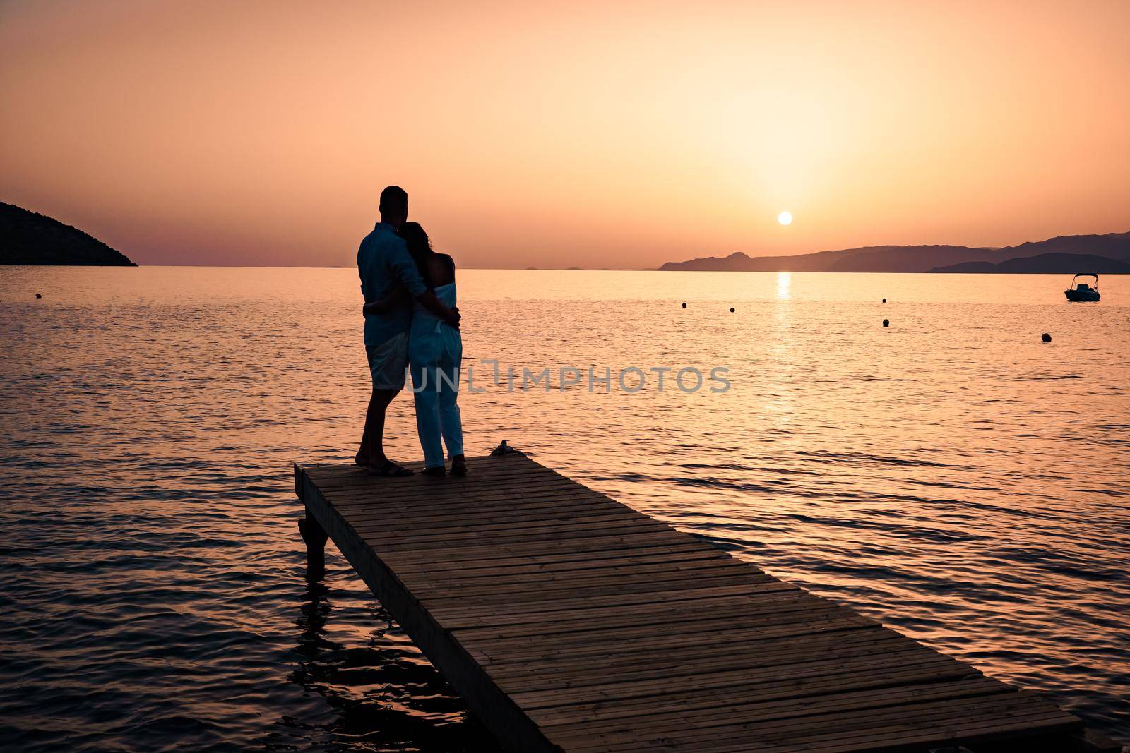 couple seated on a wooden jetty, looking a colorful sunset on the sea , men and woman watching sunset in Crete Greece by fokkebok