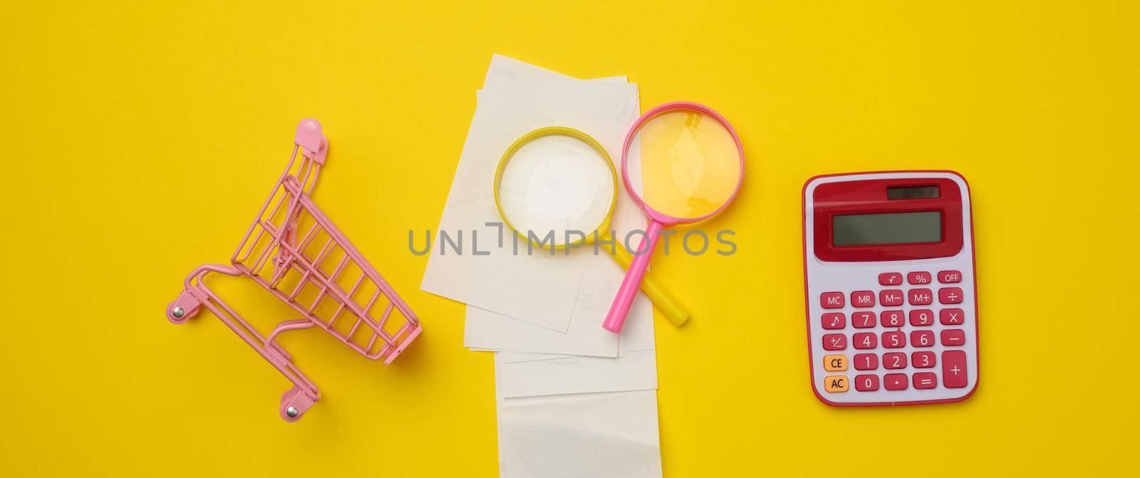 stack of paper checks, a pink plastic calculator and a magnifying glass on a yellow background. Family budget audit concept, search for savings